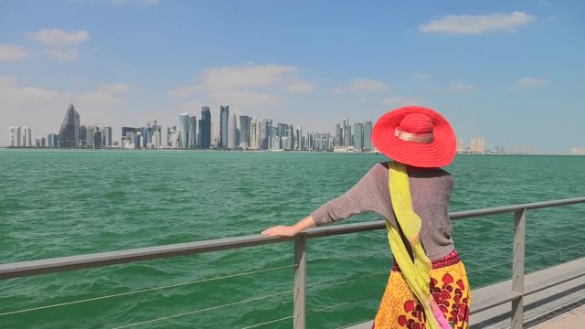 Happy woman with sunhat along the Doha Bay looking seascape and cityscape on background. Lifestyle caucasian tourist enjoys skyscrapers of Doha Downtown skyline. Qatar, Middle East travel concept.