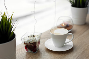 White cup of hot cappuccino on white saucer and red velvet dessert on wooden bar table next to window. Cold snowy weather outside