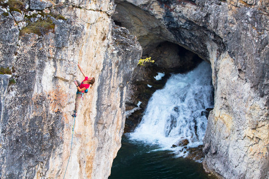 A woman warming up on a beautiful 5.11 sport climb at Natural Bridge State Park. The iconic Natural Bridge Falls is pictured in the background. Normally 100 feet tall, during lower flow times in the summer, fall and early winter the  waters from the Main Boulder River flow into a sinkhole above the falls and come out halfway down the limestone face.