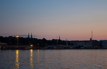 Fototapeta na wymiar View of the silhouettes of the city of Helsinki with lights on the embankment and the spiers of cathedrals from the Gulf of Finland at dusk on a bright summer night in the capital of Finland. Backgrou