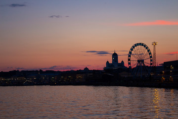 Obraz premium View of the embankment of Helsinki, the Ferris wheel, lights and the silhouette of the Cathedral of St. Nicholas from the Gulf of Finland at dusk on a bright summer night in the capital of Finland.