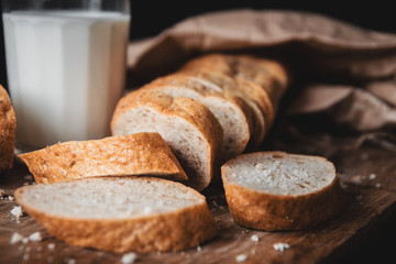 Healthy food. Long loaf of rural bread with two cut-off pieces lie on a wooden chopping board and a glass of fresh milk. Dark background.