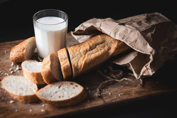 Healthy food. Long loaf of rural bread with two cut-off pieces lie on a wooden chopping board and a glass of fresh milk. Dark background.