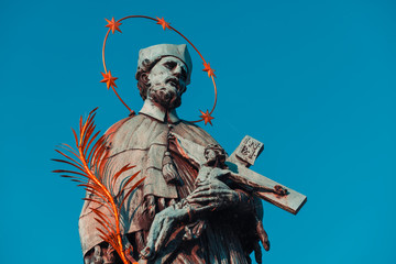 Naklejka premium Baroque Statue of Saint John Nepomuk with golden star halo holding crucifix with Jesus on Charles Bridge. Prague, Czech Republic. Orange and teal mood