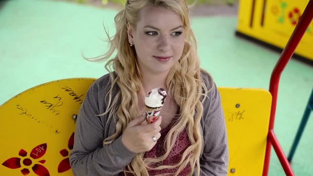 Young lovely blonde girl at swings eating and licking ice cream at cloudy weather