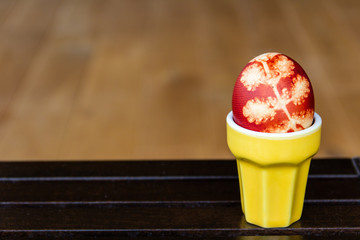 One Easter egg dyed with onion peels with a pattern of plants and leaves, put in yellow ceramic cup, on dark wooden table, on blurred wooden background. Close-up with copy space.