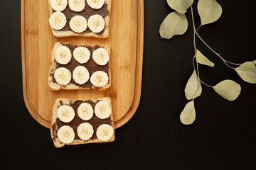Three banana white bread toasts spread with chocolate butter that lie on a chopping board with a sprig of leaves on a dark background. top view with area for text