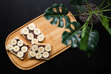 Three banana white bread toasts spread with chocolate butter that lie on a chopping board with a sprig of leaves on a dark background. top view with area for text