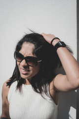 An Indian origin woman in her 40s, plays with her hair in front of a white wall, wearing sunglasses on a hot sunny day. 