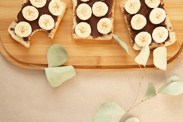 Three banana white bread toasts smeared with chocolate butter that lie on a cutting board with a sprig of leaves on craft paper background. top view with area for text