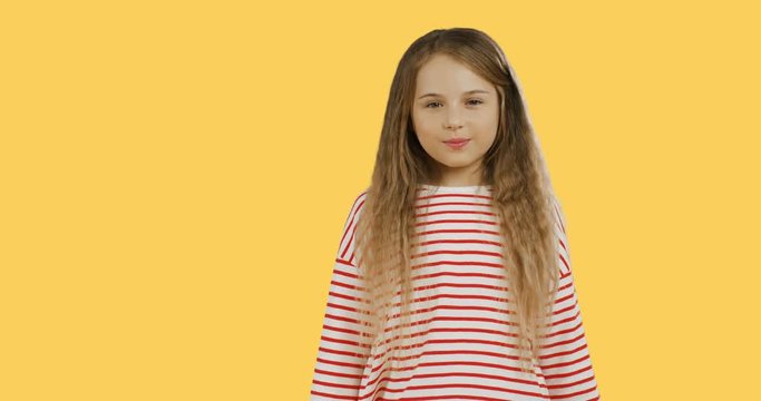 Funny small schoolgirl with long hair standing on the chroma key background, showing her tangue to the camera and crossing her hands. Yellow wall on the back.
