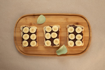 Three banana white bread toasts smeared with chocolate butter that lie on a cutting board with a sprig of leaves on craft paper background. top view with area for text