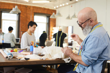 Mature grey-bearded man in shirt sitting by table and sewing white dress or other item for client in workshop