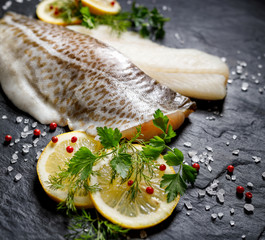 Fresh raw cod fillet with addition of herbs and lemon slices on black stone background, close-up