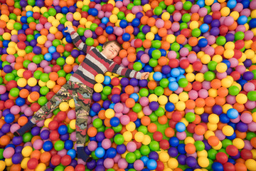 Emotional cheerful little child having fun in entertainment game center. Happy baby boy lying in pool filled with lot of colorful foam rubber balls in children's entertainment complex. Copy space