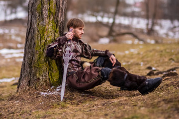 Costume Play. Caucasian Knight with Sword Sitting Near Tree Against Nature River Background.