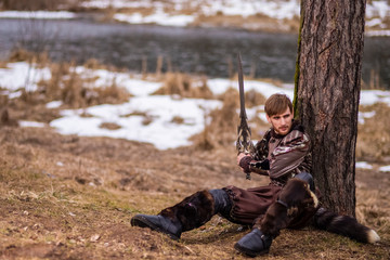 Costume Play. Caucasian Knight with Sword Sitting Near Tree Against Nature River Background.