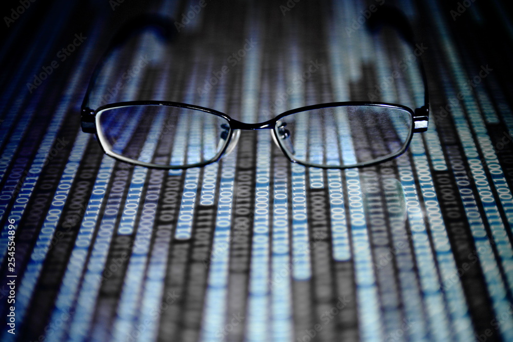 Wall mural reading data in digital age. glasses and flowing binary code matrix. shallow depth of field image of glasses on display panel. multiple lines of blue code moving under glasses.