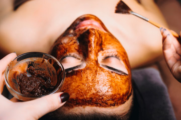 Hands of a cosmetologist holding a chocolate mask and a brush doing a chocolate mask to a woman.