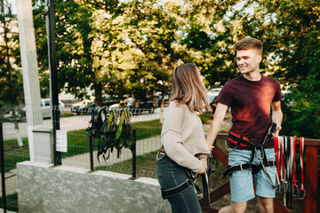 Beautiful young caucasian couple with ropeway equipment on them having fun because of adrenaline looking one tho another smiling outside on rope slide platform while dating.