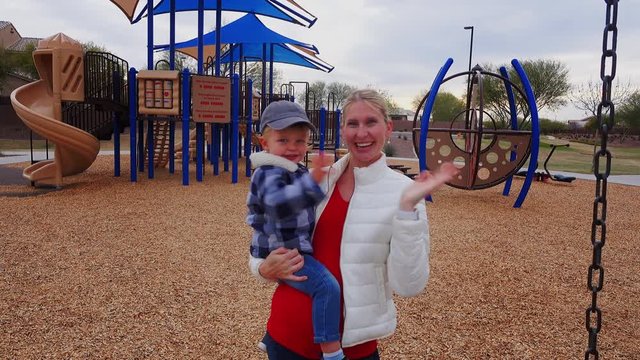 A young mother holds her son and waves to the camera in a typical Arizona neighborhood playground on a chilly winter day. Phoenix suburbs.  	