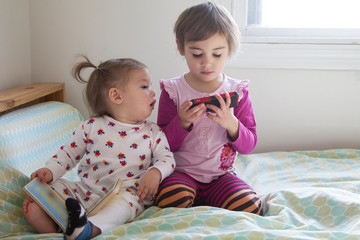 4-year-old girl watching cell phone and 1-year-old girl reading book