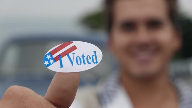 Young man lifts up a generic "I Voted" sticker in the frame, smiles in back.