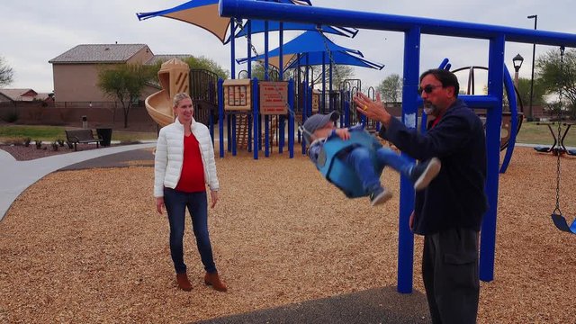 A mother and grandfather play with their child on a swingset in a typical Arizona neighborhood playground. Phoenix suburbs.  	