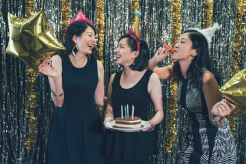 Happy girl friends celebrating birthday with cake and gold star balloons blowing whistle toy. group of cheerful ladies having fun at 20s party in nightclub. charming women wearing glamorous dresses.