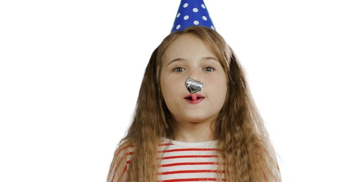 Close up of the funny cute small schoolgirl with a long hair in the birthday party hat and blowing in the horn. White wall back.