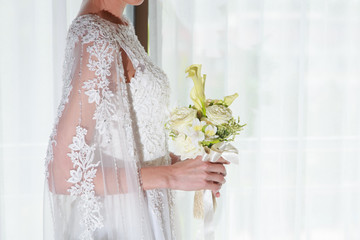 Portrait of the beautiful bride holding the bouquet near the window indoors