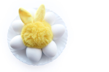 bright yellow toy bunny on a plate with white eggs on a white background. concept easter. copy space..