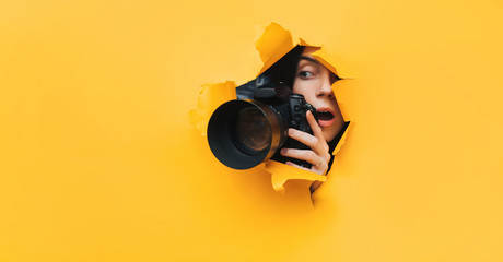 A paparazzi girl with a camera looks out from cover and looks at what is happening with her mouth open. Yellow paper, torn hole. Tabloid press. Copy space. - obrazy, fototapety, plakaty