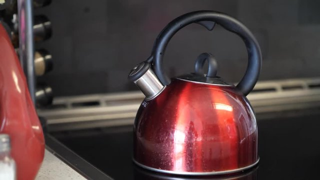 A close up of a used shiny red tea kettle with round black handle and chrome spout on a stove blowing out steam and making a shrieking whistle noise indicating its hot.