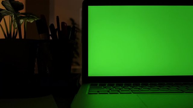 Laptop Computer Showing Green Chroma Key Screen Stands on a Desk in the Living Room. In the Background Cozy Living Room in the Evening with Warm Lights on. Dolly shot Left to Right. 4K