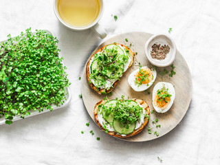 Breakfast, snack, brunch - cream cheese, cucumber, micro greens sandwiches, boiled egg and green tea on a light background, top view