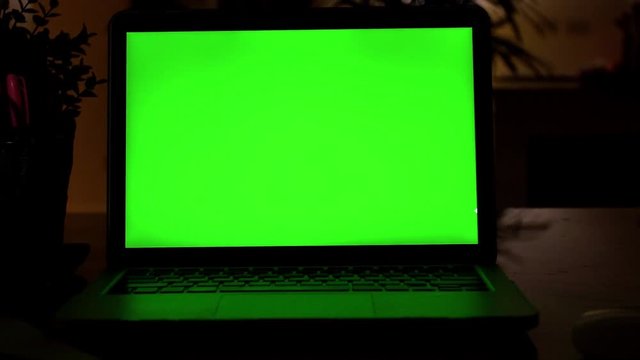 Laptop Computer Showing Green Chroma Key Screen Stands on a Desk in the Living Room. In the Background Cozy Living Room in the Evening with Warm Lights on. Dolly shot Right to Left. 4K