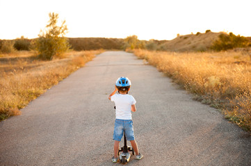 active healthy little boy in helmet and denim shorts standing on empty road with scooter in sunset