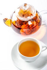 A Cup of fresh tea on a saucer, behind the Tea in a glass teapot with a blooming large flower. Exotic green tea on white background