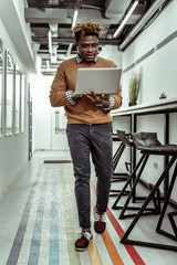 Dark-skinned focused man with funky hairstyle moving around with laptop