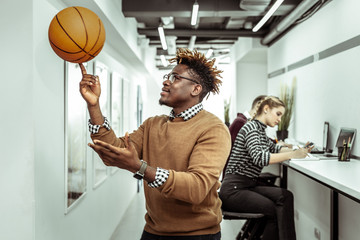 Active contented guy with dreadlocks rolling basketball ball on one finger