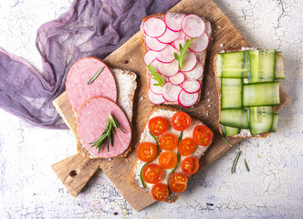 Set of various sandwiches with radish, cucumber, tomatoes and sausage, seasoned with arugula and rosemary on a rustic desk on a light background top view