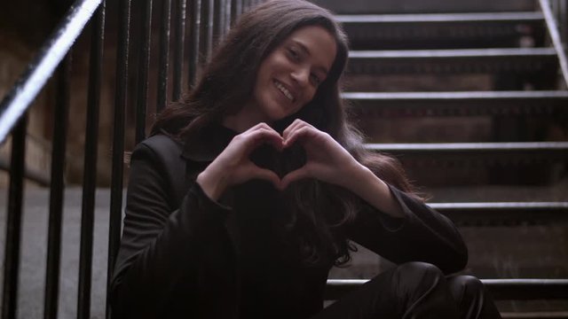 Medium shot of an attractive woman making a heart symbol both hands while sitting outdoors on some stairs