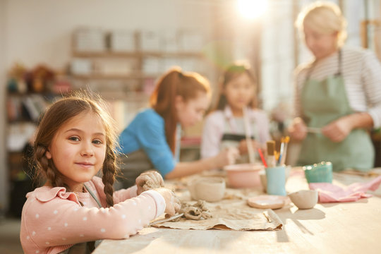 Portrait of cute little girl looking at camera while enjoying pottery class with group of children, copy space