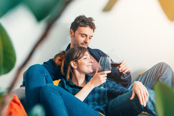 young couple sitting on sofa at home, toasting and smiling - home life, living together, and domestic partnership concept