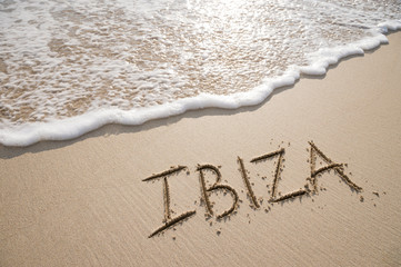 Simple Ibiza Spanish holiday getaway message written in smooth sand with an incoming wave on a tropical beach - obrazy, fototapety, plakaty