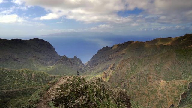Panoramic view of a landscape of mountains and two tourists standing on the cliffs and taking photos. The Atlantic ocean and the island of La Gomera in the background