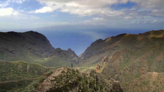 Panoramic view of a landscape of mountains and two tourists standing on the cliffs and taking photos. The Atlantic ocean and the island of La Gomera in the background. HD cropped edit