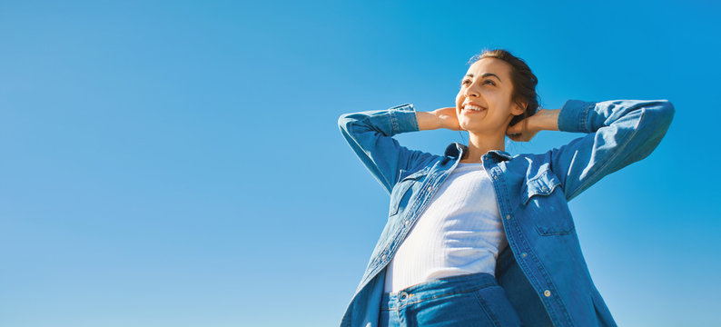 bottom view of a young smiling attractive woman in jeans clothes at sunny day on the blue sky background. joyful woman posing outdoors