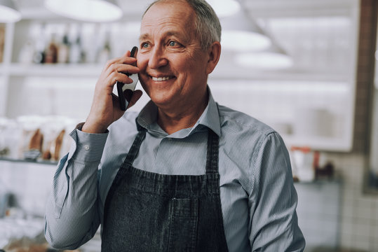 Joyful senior man in apron talking on cellphone